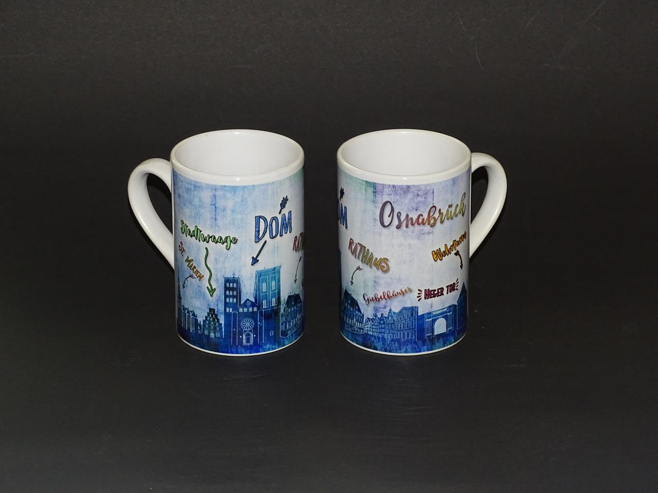 Auf dem Bild sind zwei Tassen mit dem Aufdruck "Tasse Slim Vintage Osnabrück" zu sehen. Die Tassen zeigen eine blaue Skyline und Schriftzüge von Sehenswürdigkeiten aus Osnabrück.