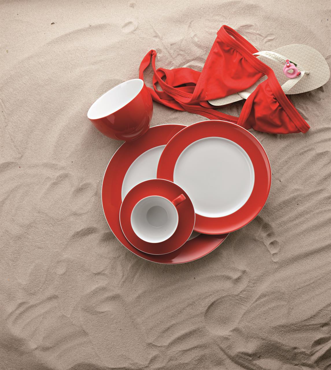 Das Bild zeigt einen Speiseteller, eine Tasse und weiteres Geschirr der Serie "Sunny Day / New Red" auf sandigem Untergrund. Daneben liegen ein rotes Bikini-Oberteil und weiße Flip-Flops.