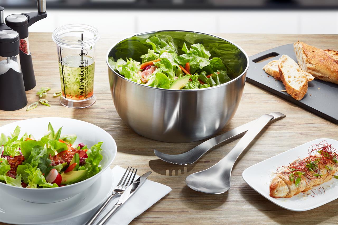 Auf dem Bild ist die Salatschleuder SPEEDWING® aus Edelstahl zu sehen, gefüllt mit frischem Salat. Daneben stehen Salatbesteck, ein Schälchen mit Dressing und geschnittenes Baguette auf einem Tisch.