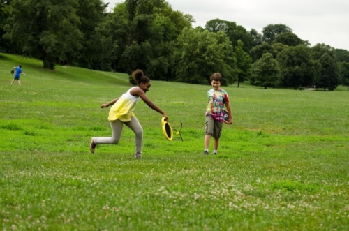 Zwei Kinder spielen zusammen mit einem Frisbee auf einer Wiese. Im Hintergrund sind Bäume und eine weitere Person zu sehen.