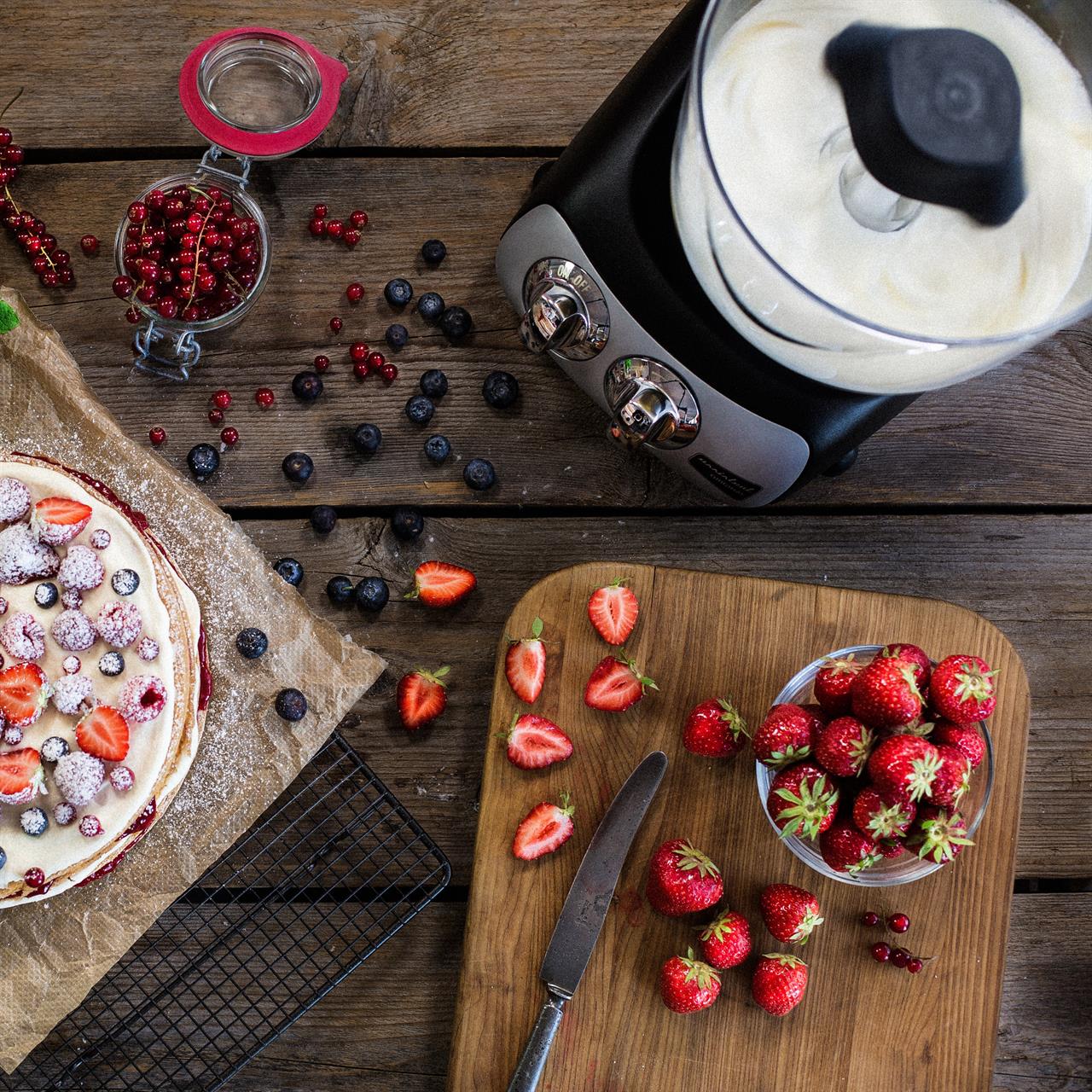 Auf dem Bild ist die Küchenmaschine Black zu sehen, die Sahne aufschlägt. Daneben liegen frische Beeren, ein Kuchen und ein Schneidebrett mit Erdbeeren auf einem Holztisch.