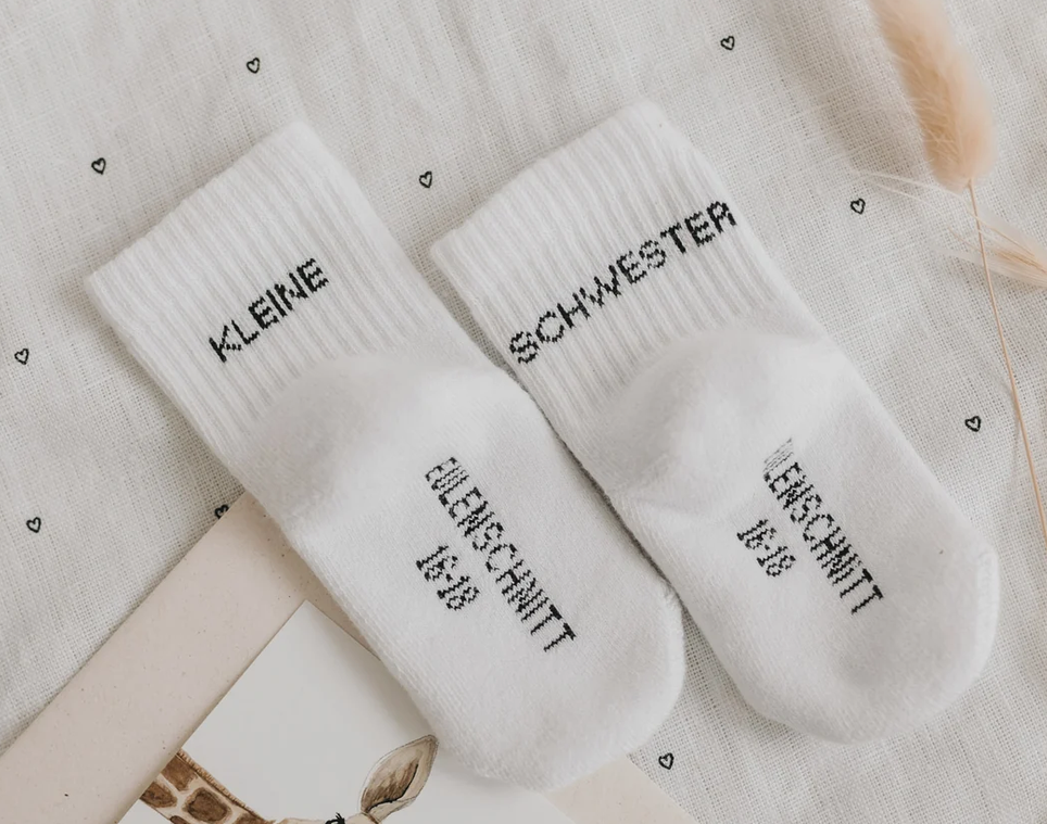 Babysocken Kleine Schwester in der Größe 16-18 sind abgebildet. Die Socken sind speziell für kleine Mädchen mit der Aufschrift „Kleine Schwester“ gestaltet.