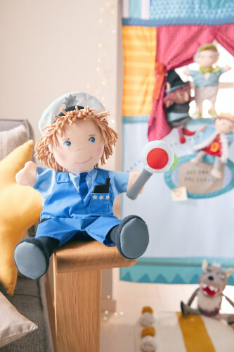 Ein Stoffpuppe in einer blauen Polizeiuniform sitzt auf einer Holzlehne. Im Hintergrund sind weitere Puppen vor einem bunten Theater-Vorhang zu sehen.