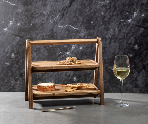 Der Servierturm "Gather" aus Akazienholz ist zweistöckig und eignet sich ideal zum stilvollen Anrichten von Snacks. Auf dem unteren Brett liegen Käse und Cracker, auf dem oberen Walnüsse; daneben steht ein Glas Weißwein.
