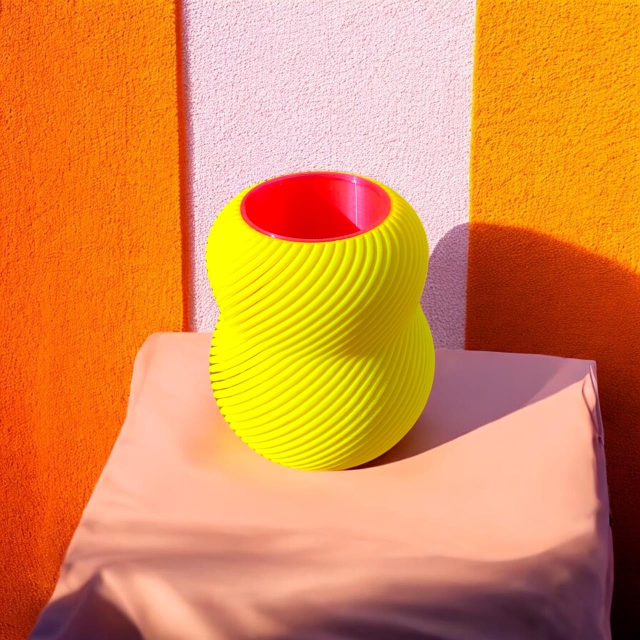 Die Abbildung zeigt eine Vase mit leuchtend gelber spiralförmiger Oberfläche und rotem Inneren. Sie steht auf einem beigefarbenen Tuch vor einer Wand in Orange- und Rosatönen.