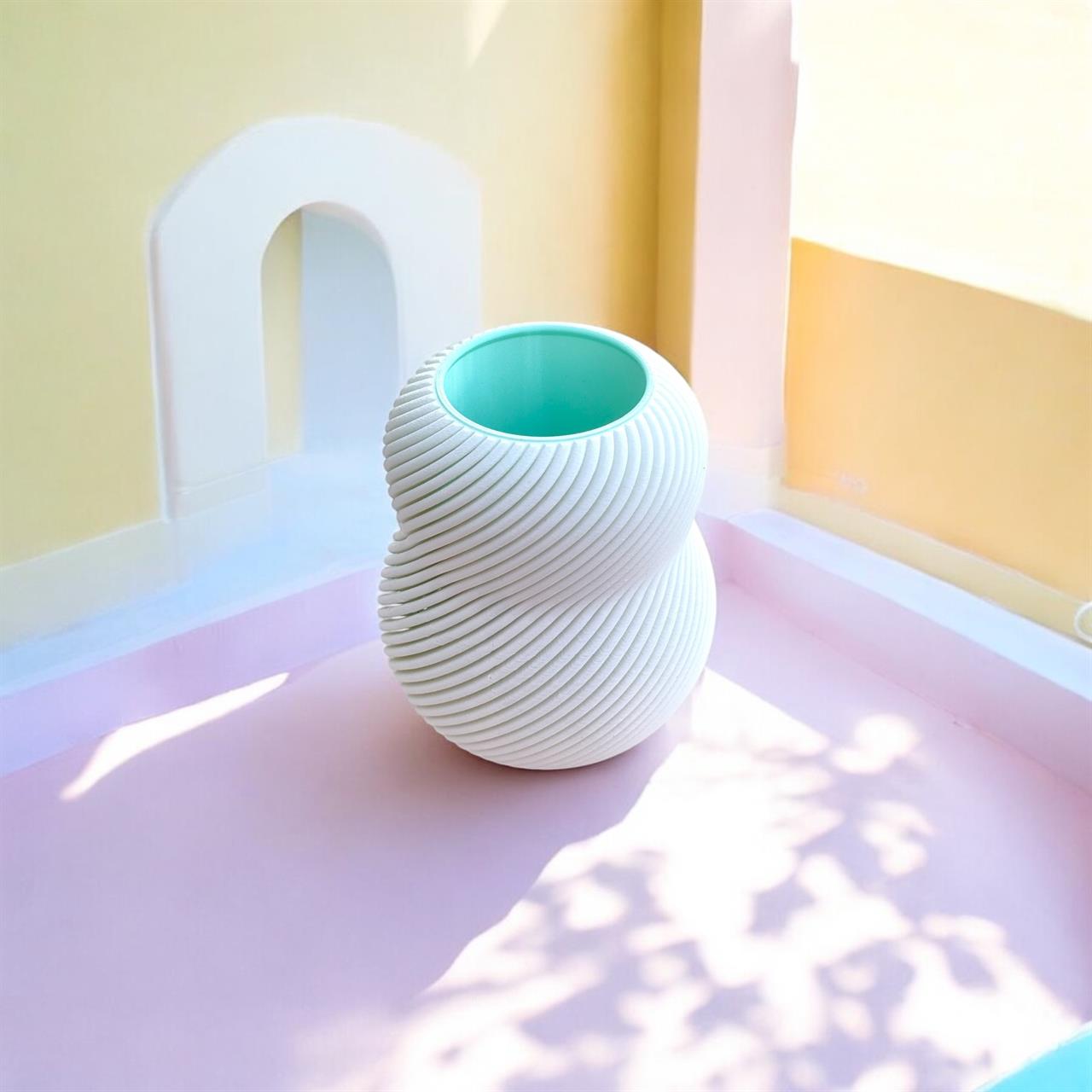 Das Bild zeigt die Vase "Vase Bloom 100% White" mit einem modernen, gedrehten Design. Sie steht in einem hellen, pastellfarbenen Raum mit weichem Lichteinfall.