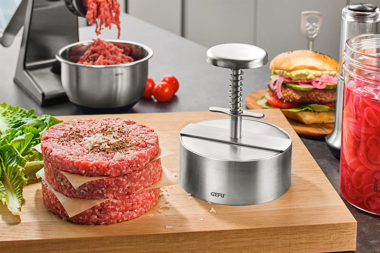Die Profi-Hamburgerpresse PÄTTI ist auf einem Küchentisch neben frisch geformten Burgerpatties zu sehen. Im Hintergrund stehen Zutaten und ein fertiger Burger bereit.