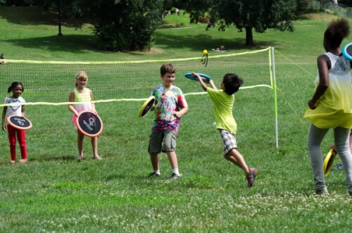 Mehrere Kinder spielen mit Wurfscheiben auf einer Wiese. Sie haben ein Netz aufgespannt und scheinen viel Spaß zu haben.