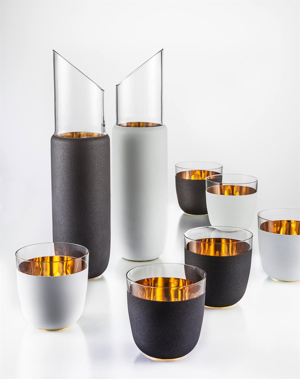 Auf dem Bild sind zwei elegante Karaffen und mehrere passende Trinkgläser zu sehen, die in Schwarz und Weiß gehalten sind. Die Gläser und Karaffen haben eine matte Außenseite und einen glänzenden, goldfarbenen Innenbereich.