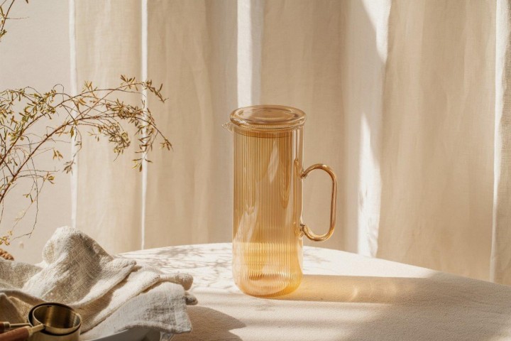 Auf dem Bild ist eine goldene Glaskaraffe mit Henkel und Deckel zu sehen. Sie steht auf einem Tisch in warmem Sonnenlicht vor einem hellen Vorhang.