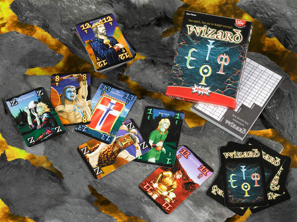Das Bild zeigt das Kartenspiel "Wizard" mit mehreren ausgebreiteten Spielkarten und der Spieleschachtel. Im Hintergrund sind außerdem ein Block mit Wertungsbögen und weitere Kartenstapel zu sehen.