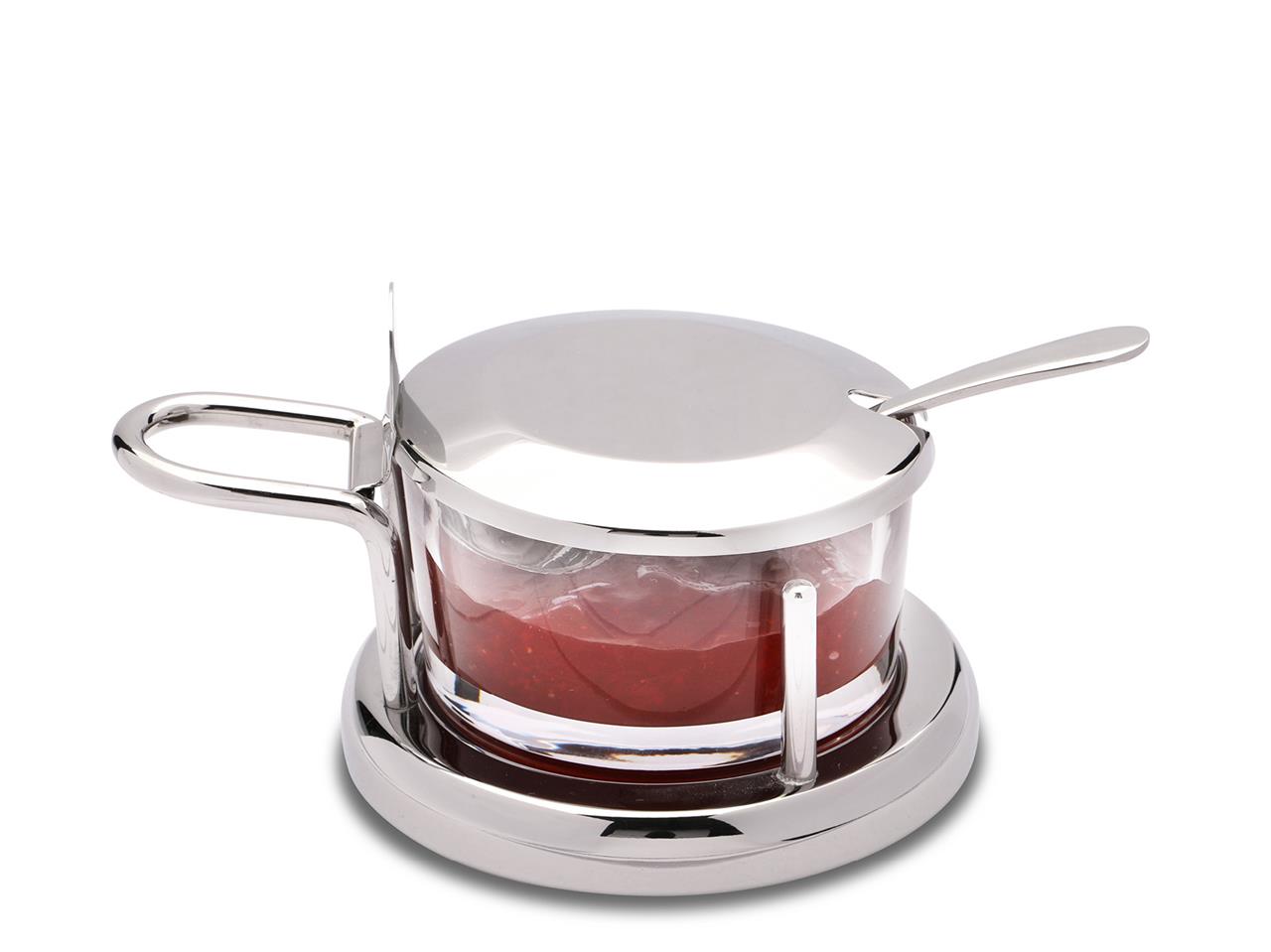 Das Bild zeigt eine Parmesan- oder Marmeladendose mit Löffel. Die Dose besteht aus Glas und Metall und steht auf einem passenden Untersetzer.