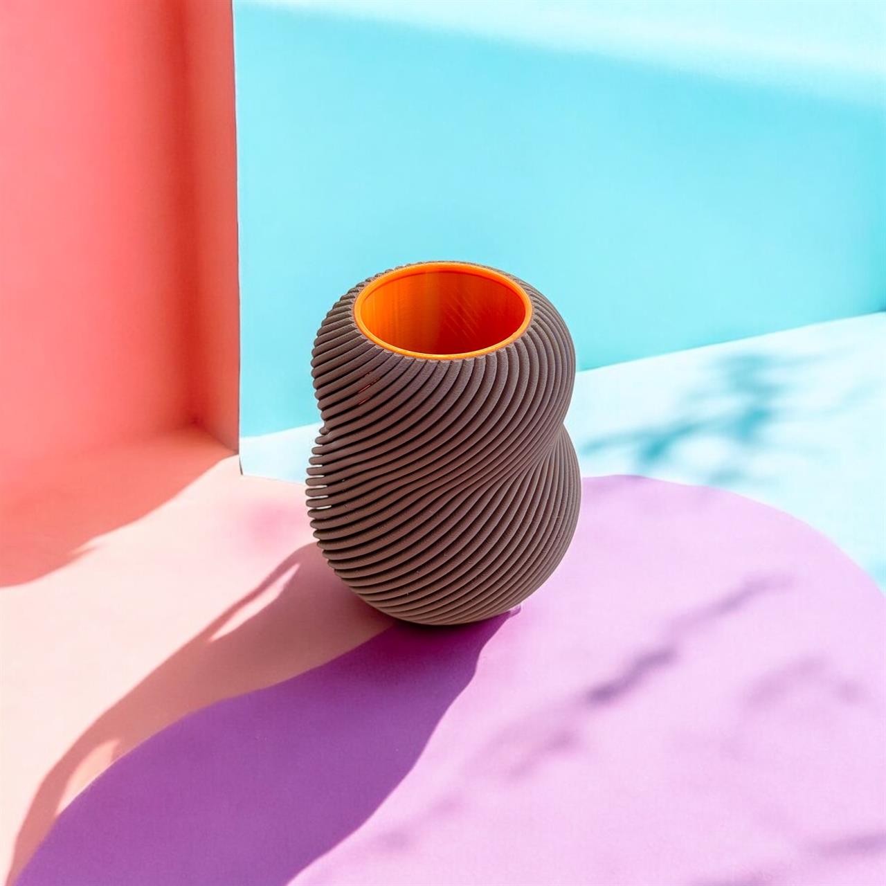 Die Vase "Vase Bloom 100% Chocolate" steht auf einer farbenfrohen Oberfläche mit rosa, lila und blauen Tönen. Sie hat eine gewundene, strukturierte Außenseite in Schokoladenbraun und ein leuchtend orangefarbenes Inneres.