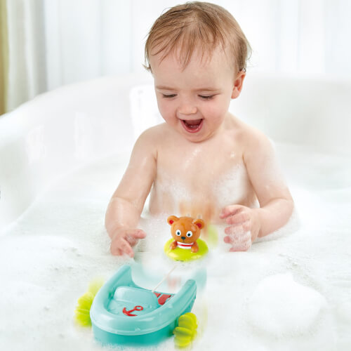 Ein Baby sitzt fröhlich in einer Badewanne voller Schaumbad. Es spielt mit Badespielzeug, darunter ein kleines Boot und eine Figur.