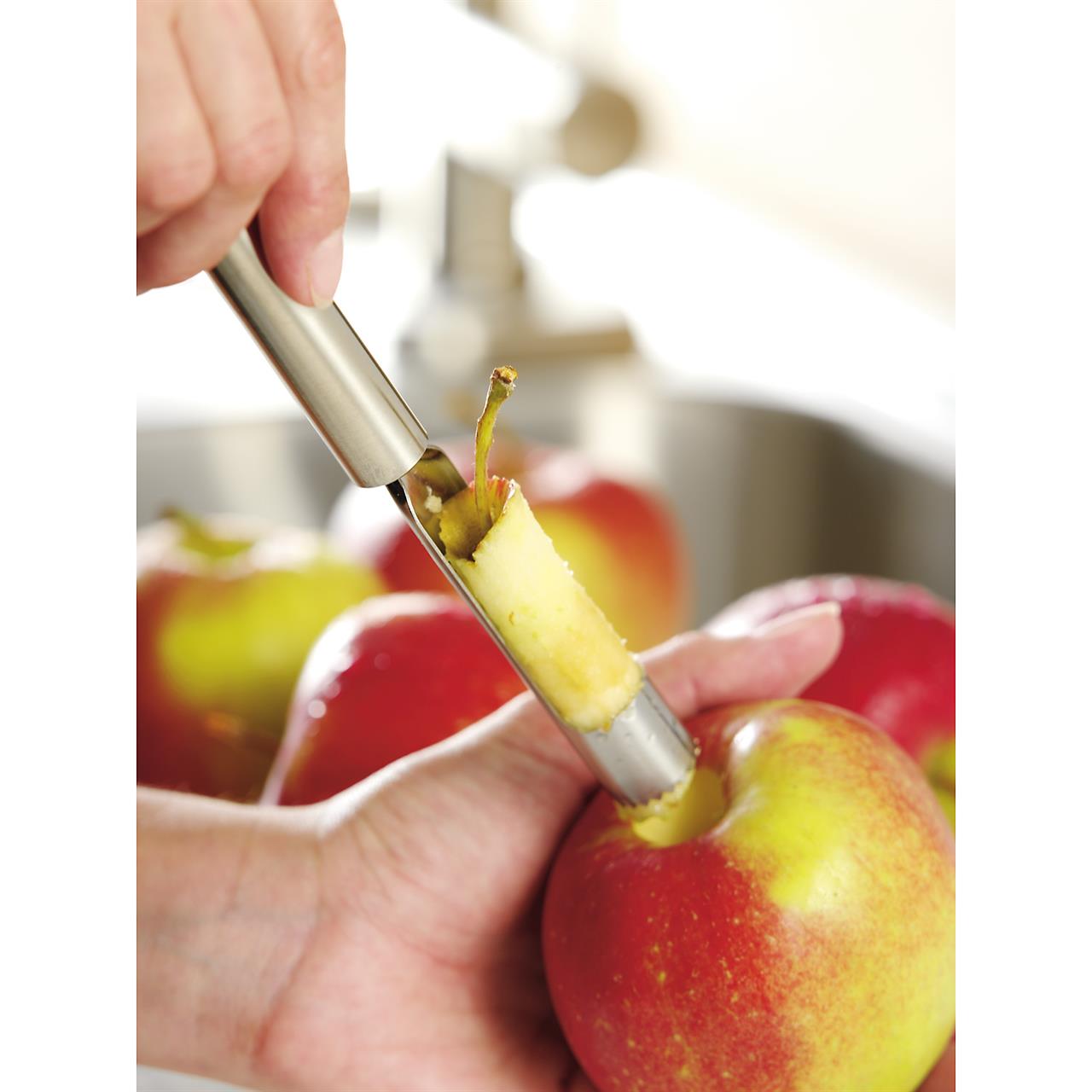 Eine Hand benutzt den Apfelausstecher Profi Plus, um das Kerngehäuse aus einem Apfel zu entfernen. Im Hintergrund sind weitere Äpfel zu sehen.