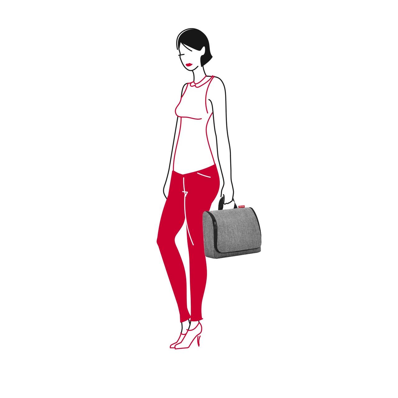 In dem Bild hält eine Frau die "Toiletbag XL twist silver Kosmetiktasche" in der Hand. Sie trägt rote Hosen, ein ärmelloses Oberteil und steht seitlich zum Betrachter.
