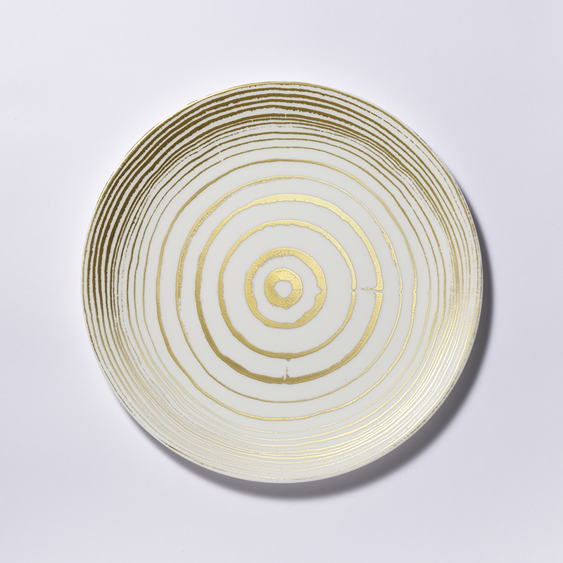 Der Teller ist rund und hat einen Durchmesser von 28 cm. Er besteht aus Fine Bone China und ist mit goldenen, ringförmigen Linien verziert, die an Jahresringe eines Baumes erinnern.