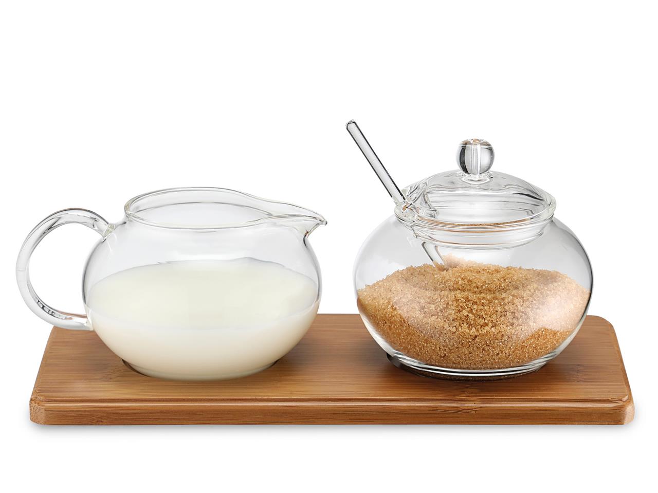 Das Bild zeigt ein Zucker-/Sahneset aus Glas, bestehend aus einem Milchkännchen und einer Zuckerdose mit Löffel. Beide Gefäße stehen auf einem rechteckigen Tablett aus Holz.
