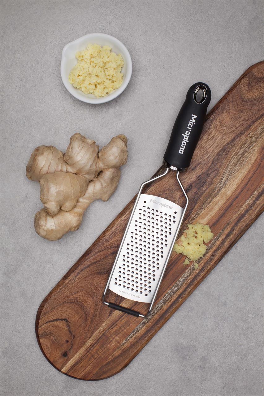 Das Bild zeigt eine grobe Reibe mit dem Namen "Reibe grob - Gourmet" auf einem Holzbrett. Daneben liegen frischer Ingwer und eine kleine Schale mit geriebenem Ingwer.