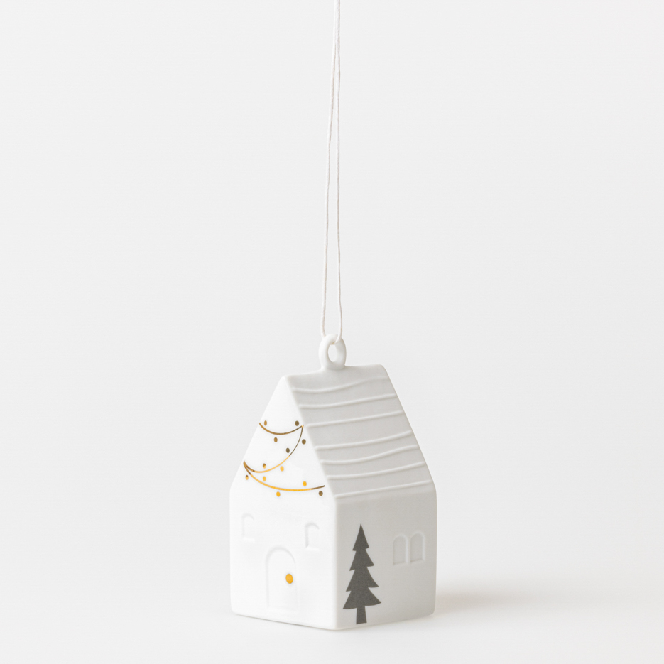 Das Bild zeigt einen weißen Weihnachtshaus-Hänger aus Keramik mit einer Größe von 5x5x10 cm. Er ist mit einer Lichterkette und einem Tannenbaum verziert und kann aufgehängt werden.