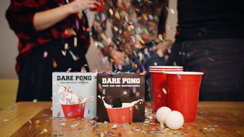 Auf dem Tisch stehen zwei Spieleboxen mit dem Namen "Dare Pong", rote Becher und zwei Tischtennisbälle. Im Hintergrund werfen Menschen Konfetti in die Luft und feiern.