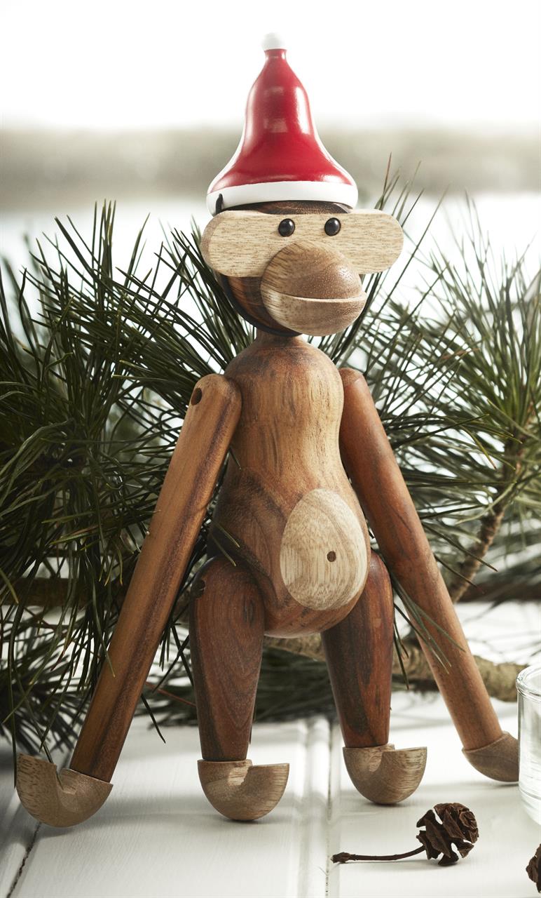 Auf dem Bild ist ein kleiner Holzaffe mit einer roten Zipfelmütze zu sehen. Er sitzt vor einigen grünen Tannenzweigen auf einer weißen Oberfläche.