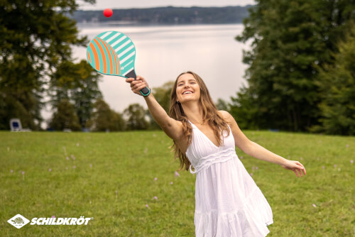 Eine Frau spielt fröhlich mit einem Schläger und einem Ball auf einer Wiese. Im Hintergrund ist ein See und Bäume zu sehen.