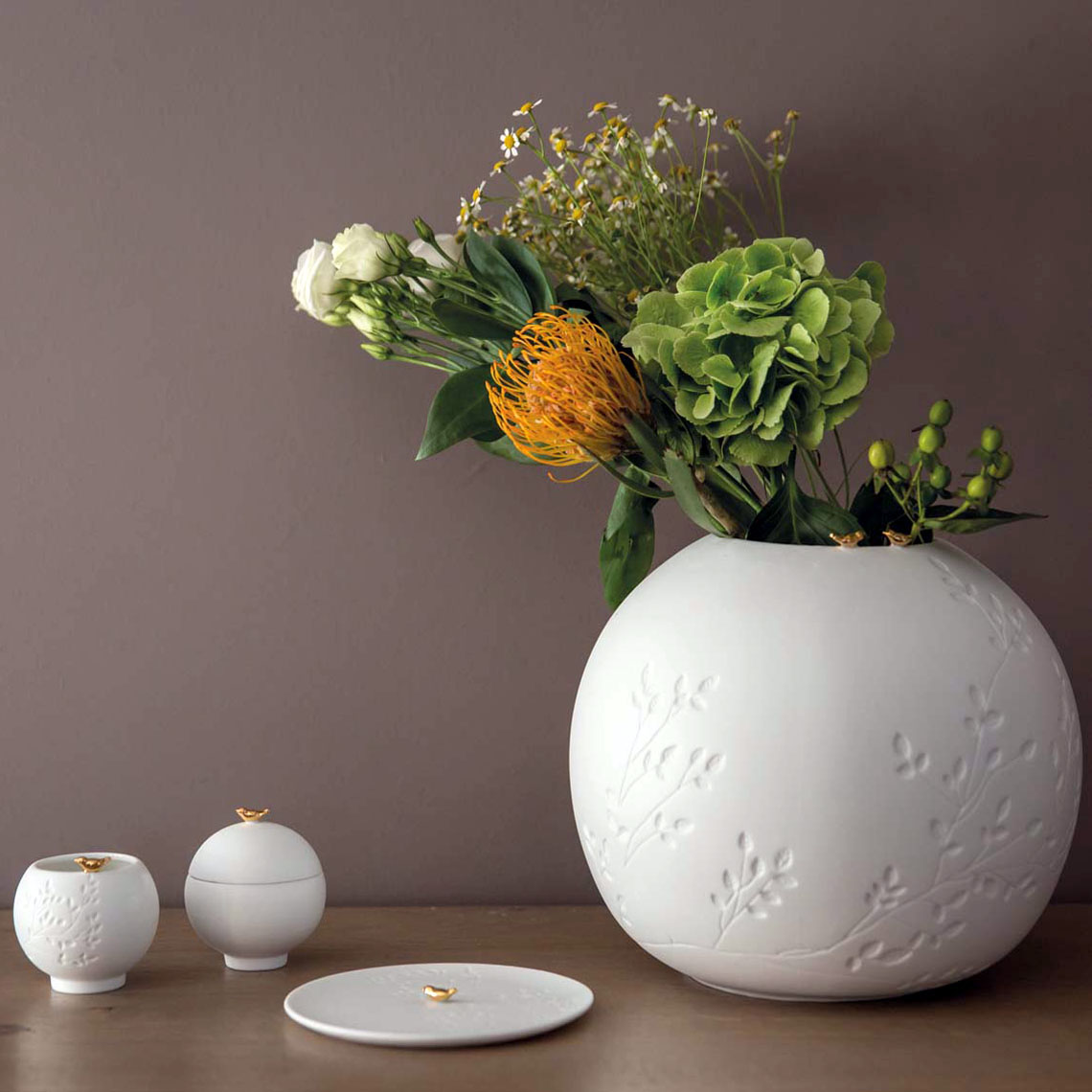 Auf dem Bild steht eine große, runde, weiße Vase mit Blumenarrangement auf einem Tisch. Daneben befinden sich drei kleinere, weiße Gefäße mit Deckeln, die kleine goldene Details haben.