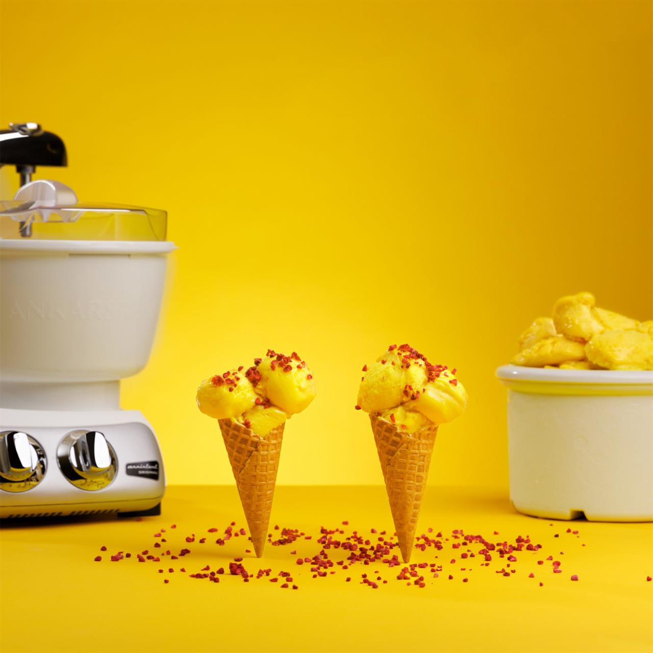Auf dem Bild ist eine Ankarsrum Eismaschine zu sehen. Davor stehen zwei Eiswaffeln mit gelbem Eis und Streuseln auf einem gelben Hintergrund.