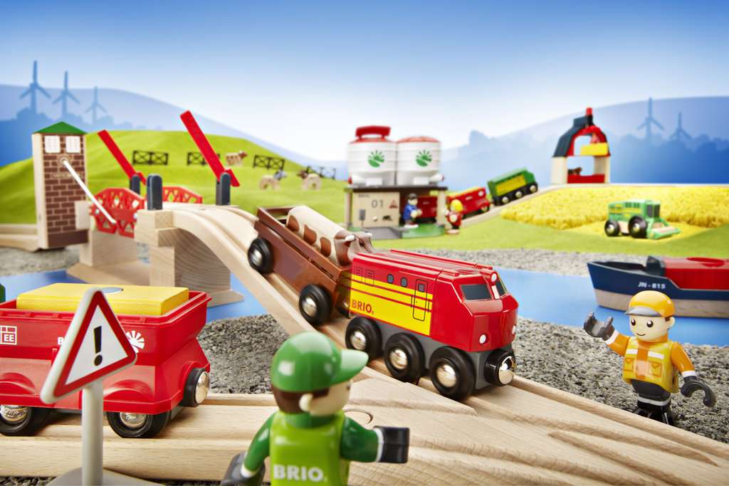 Das Bild zeigt das BRIO® Hebebrücke 33757 Spielset mit einer roten Lokomotive, Brücke und Figuren. Im Hintergrund sind weitere Spiellandschaften wie Felder, Boote und Gebäude zu sehen.