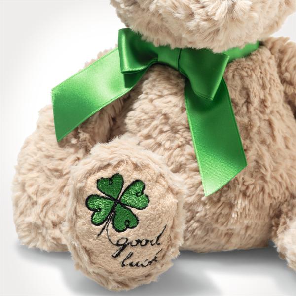 Der beigefarbene Teddybär trägt eine große grüne Schleife um den Hals. Auf seiner Pfote ist ein grünes Kleeblatt mit den Worten "good luck" aufgestickt.
