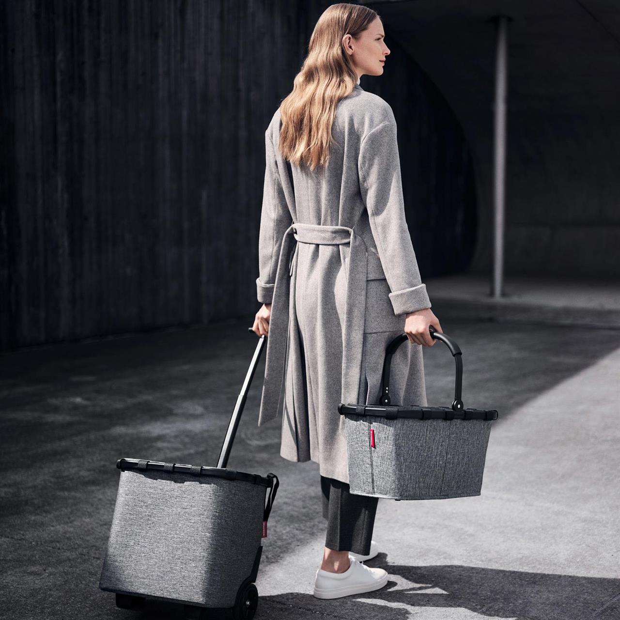 Eine Frau zieht einen grauen Carrycruiser mit Rollen und trägt einen passenden Korb in der Hand. Beide Produkte sind im modernen Design „frame twist silver“ gehalten.