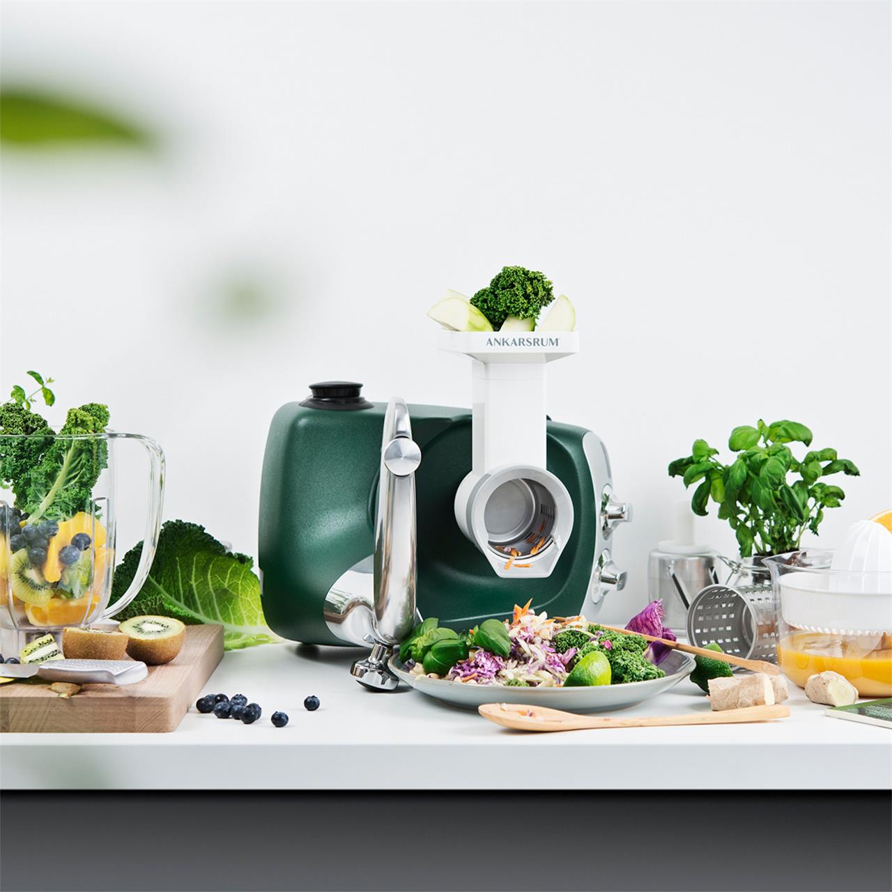 Das Bild zeigt das Produkt "Go Green Package" mit einer Küchenmaschine, die frisches Gemüse verarbeitet. Auf dem Tisch liegen verschiedene Zutaten wie Brokkoli, Kiwi, Kräuter und ein Smoothie-Glas.