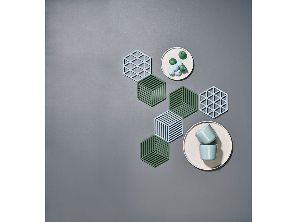 Das Bild zeigt mehrere sechseckige Topfuntersetzer in Blau und Grün, die geometrische Muster haben. Auf zwei Tellern liegen Tassen und kleine grüne Kugeln, arrangiert auf einer grauen Oberfläche.