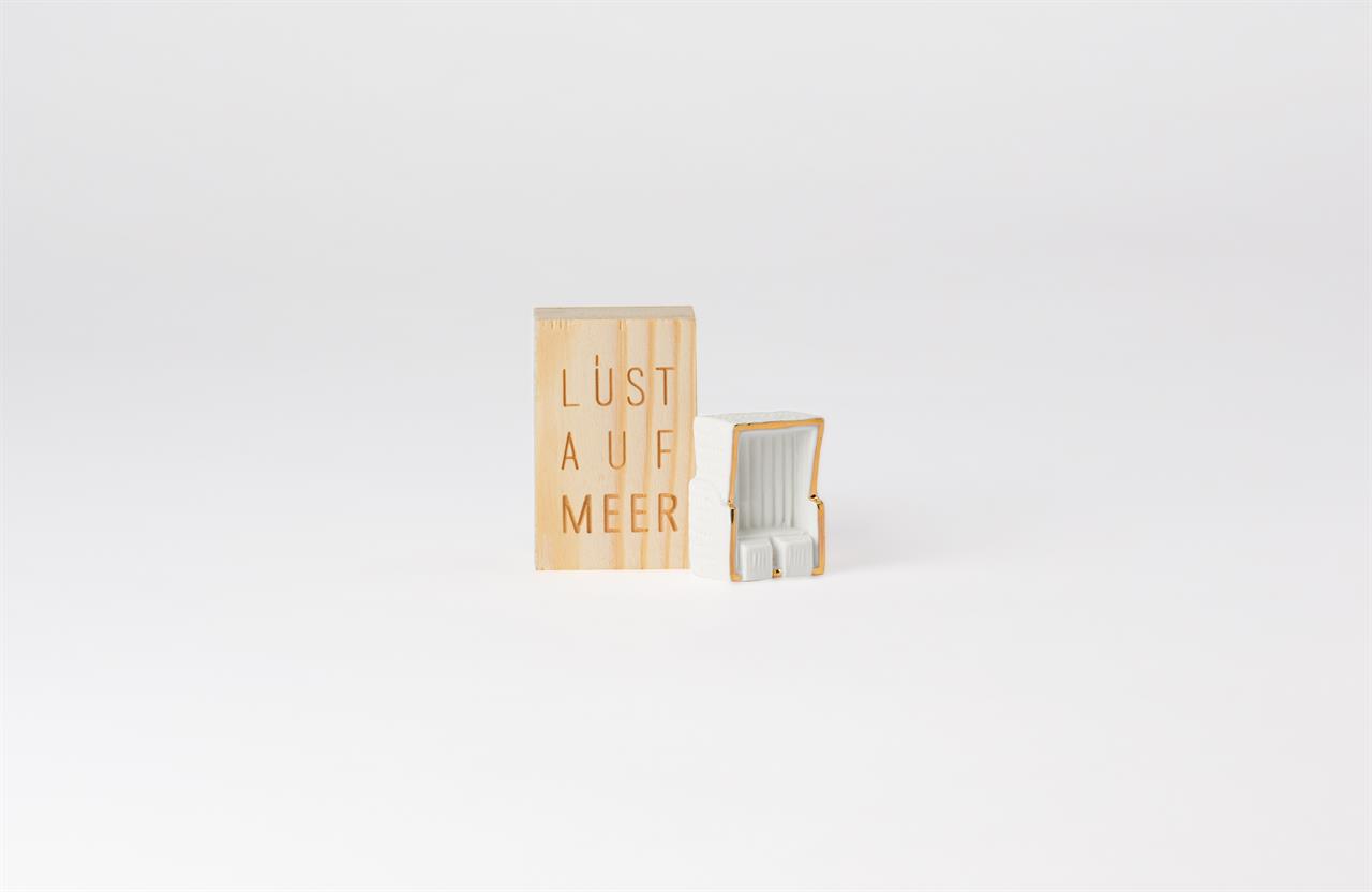 Auf dem Bild ist ein Holzgeschenk mit der Aufschrift „Lust auf Meer“ zu sehen. Daneben steht eine kleine Miniatur eines weißen Strandkorbs.