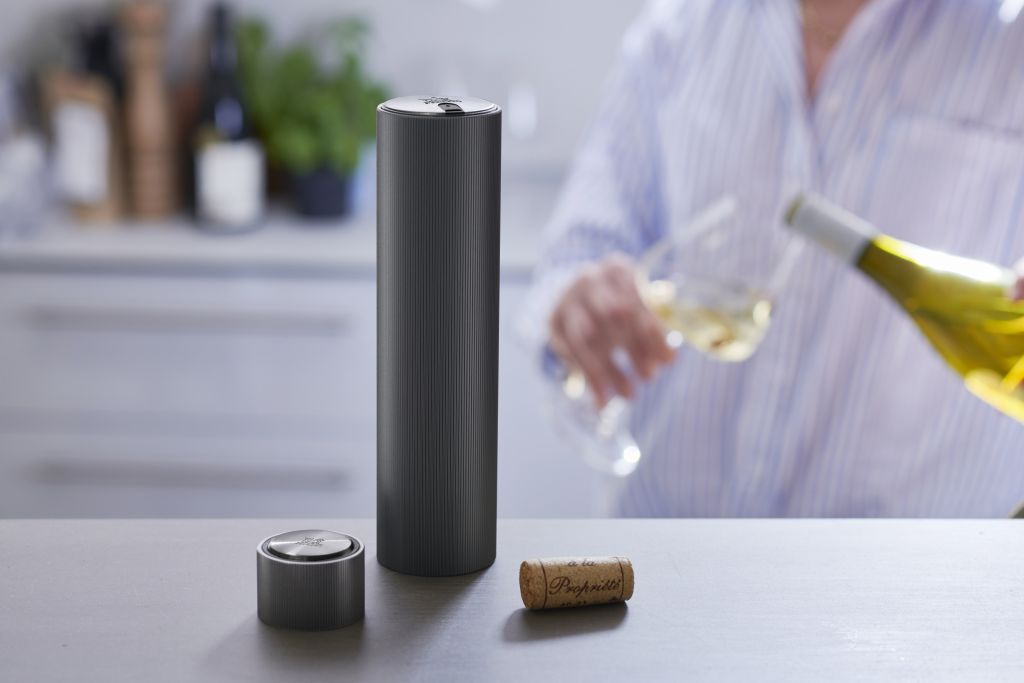 Im Vordergrund sieht man den elektrischen Korkenzieher "Carbon Line Reverse" zusammen mit einem Korken und einem Zubehörteil. Im Hintergrund wird ein Glas Weißwein eingeschenkt.