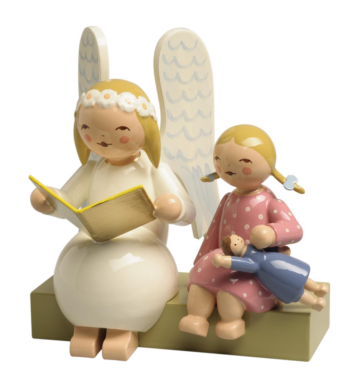 Die Figur zeigt einen Margeritenengel, der auf einer Bank sitzt und ein Buch hält. Neben ihm sitzt ein Mädchen im rosa Kleid mit einer Puppe im Arm.