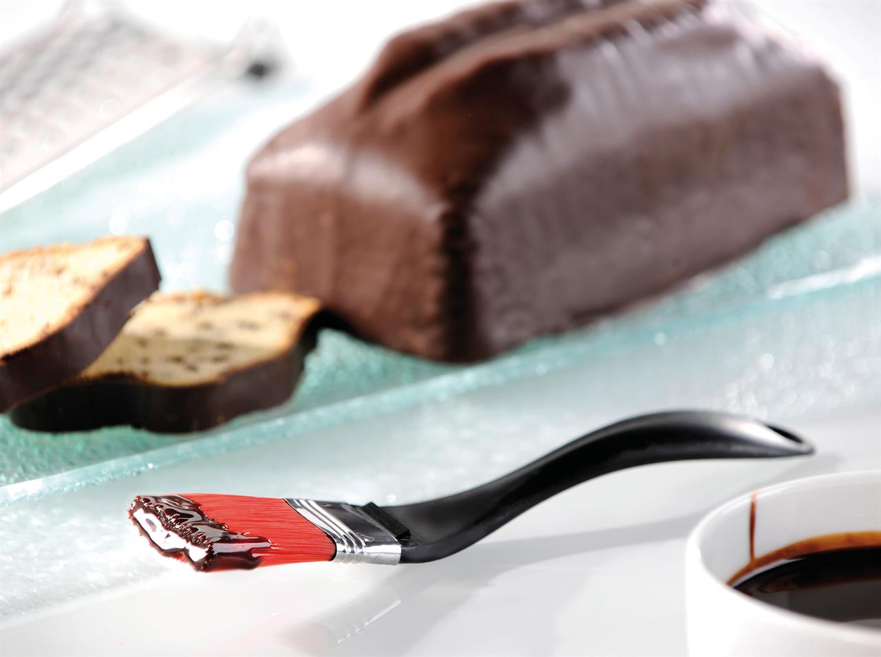 Im Vordergrund liegt der Küchenpinsel "Becky" mit rotem Borstenkopf, auf dem etwas Schokolade ist. Im Hintergrund sieht man einen Schokoladenkuchen und zwei Scheiben davon auf einem Teller.