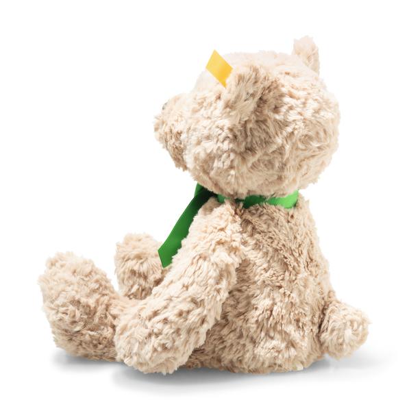 Ein beiger Teddybär sitzt seitlich und trägt ein grünes Band um den Hals. Er hat ein flauschiges Fell und einen gelben Anhänger am Ohr.
