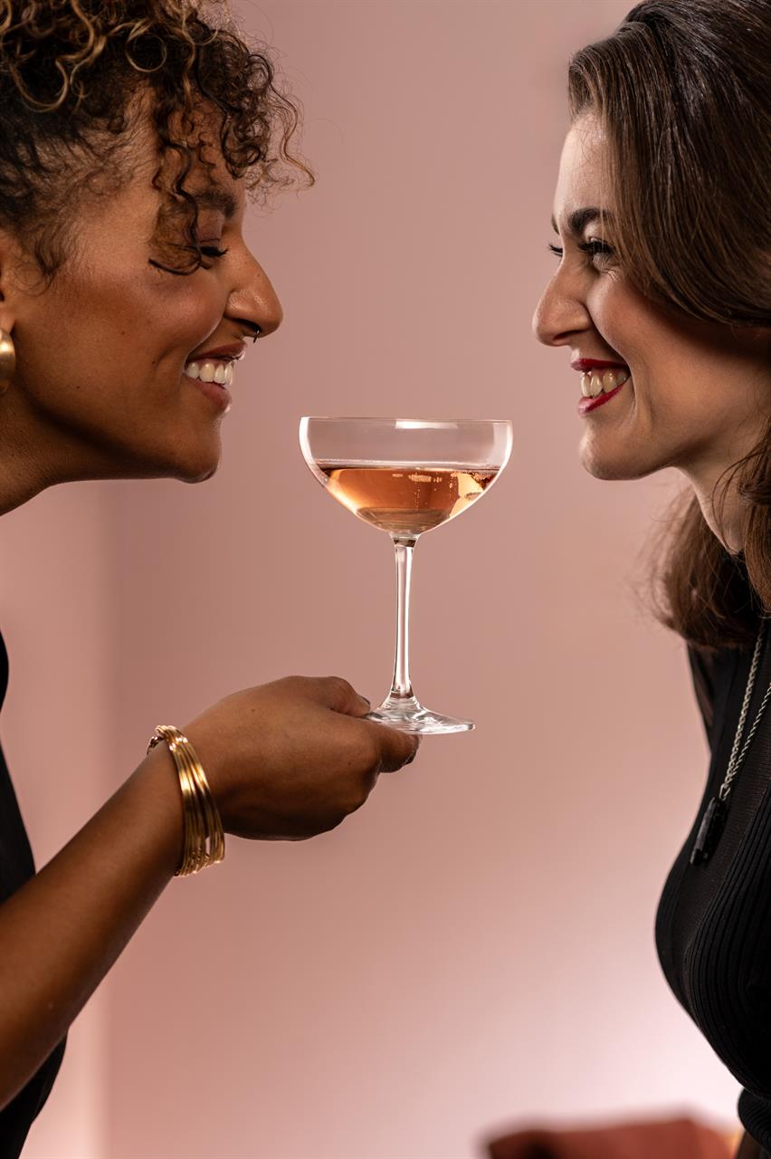 Zwei Frauen lächeln sich an und halten gemeinsam ein Sektglas. Das Glas ist ein Produkt aus der Serie "4er Sektschale Bar Special 8".
