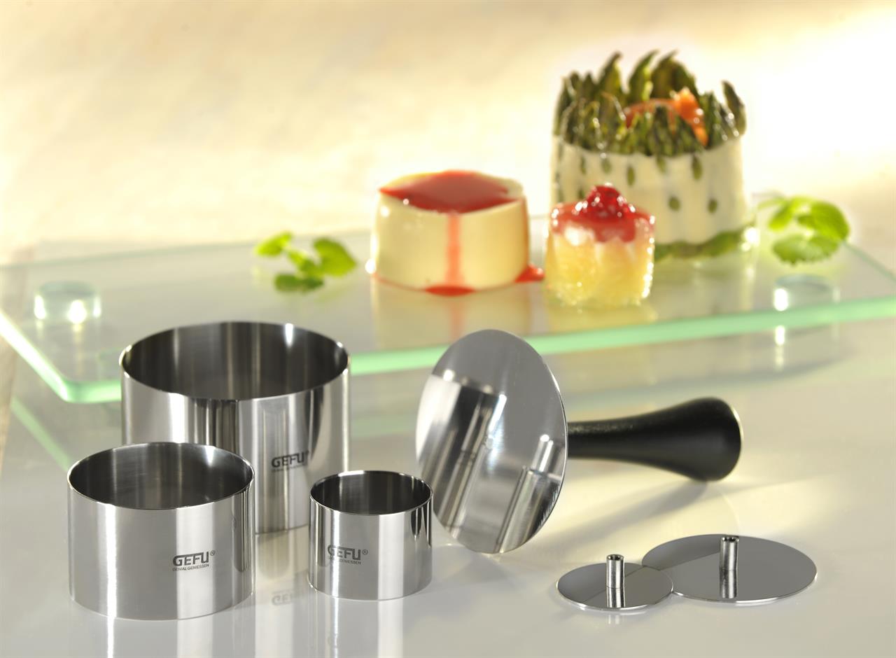 Im Vordergrund sind Profispeisenringe aus Edelstahl in verschiedenen Größen zu sehen. Im Hintergrund befinden sich kunstvoll angerichtete Speisen, die mit Hilfe der Ringe gestaltet wurden.