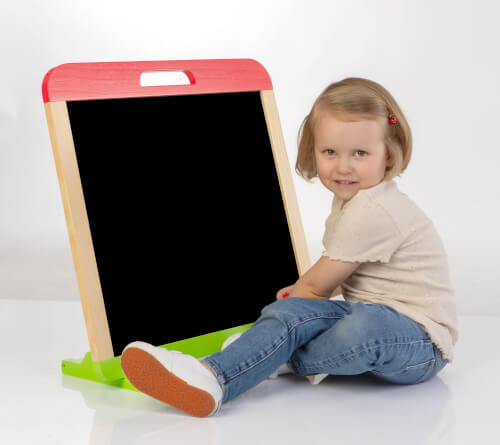 Ein kleines Mädchen sitzt auf dem Boden neben einer leeren Tafel. Sie lächelt und schaut in die Kamera.
