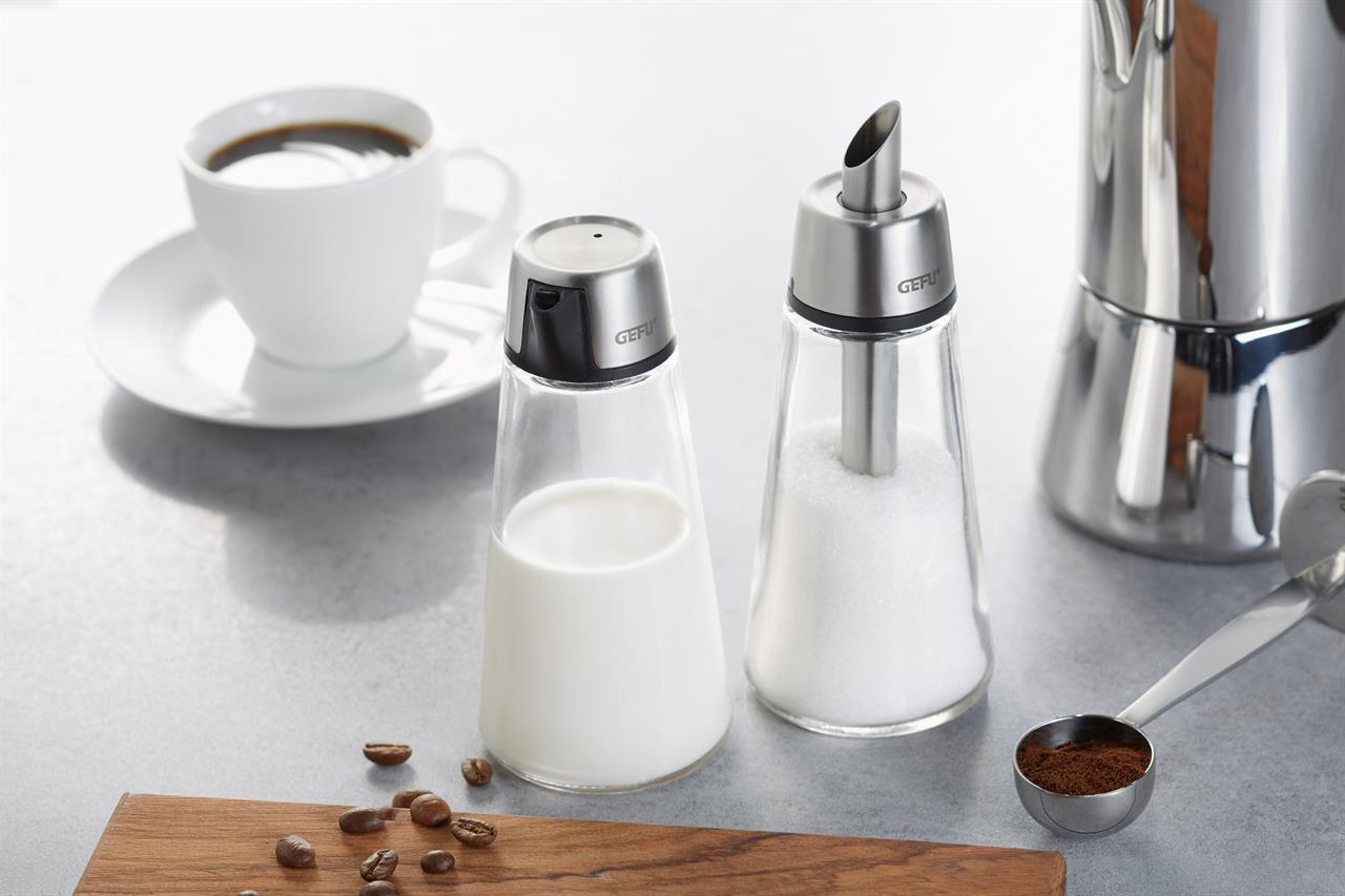 Auf dem Bild ist das Produkt "Zuckerspender Brunch" zu sehen. Es steht neben einem Milchspender, einer Tasse Kaffee und Kaffeebohnen auf einem Tisch.