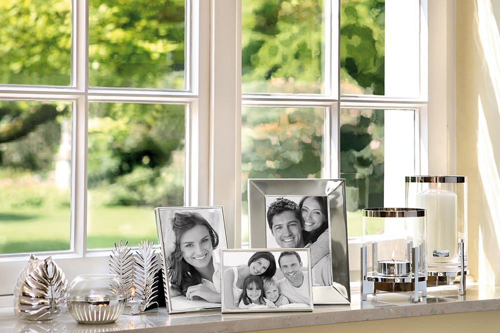 Mehrere silberne Bilderrahmen mit Fotos stehen auf einer Fensterbank. Daneben befinden sich dekorative Kerzen und glänzende Dekorationsobjekte.