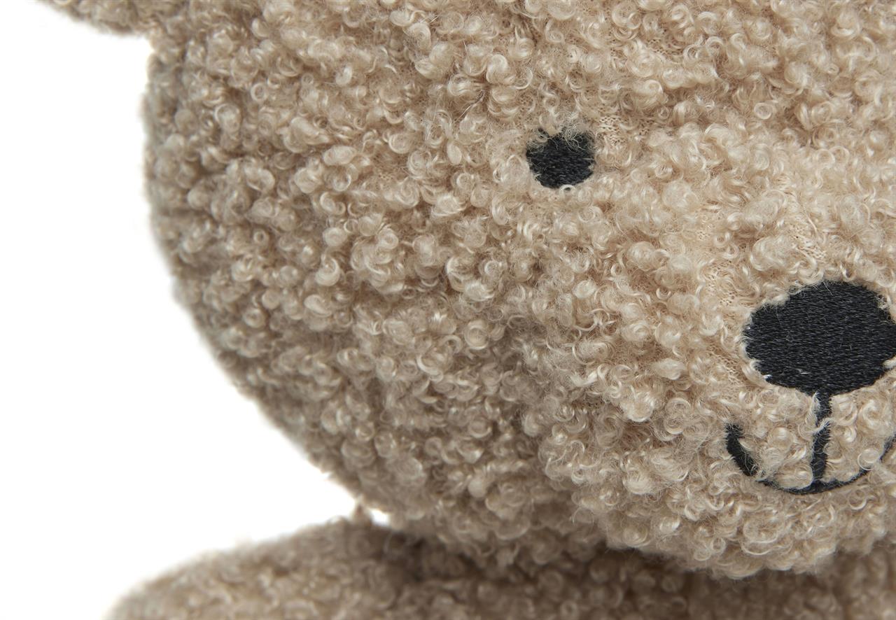 Das Bild zeigt eine Nahaufnahme eines kuscheligen Teddybären mit lockigem, beigem Fell. Man sieht deutlich das gestickte Auge und die lachende Schnauze des Teddys.