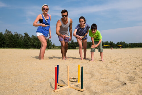 Vier Personen spielen ein Wurfspiel am Sandstrand. Sie lachen und haben Spaß zusammen.