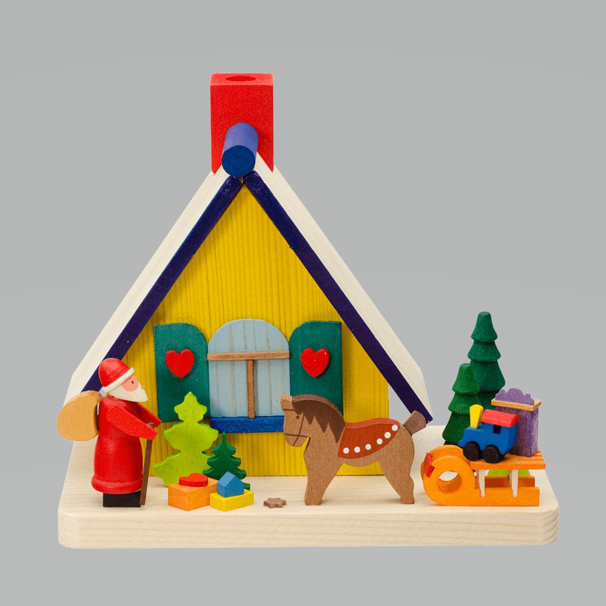 Das Bild zeigt ein buntes Räucherhaus mit einem Weihnachtsmann und verschiedenen Holzfiguren davor. Zu sehen sind ein Pferd, Geschenke, ein Baum, ein Schlitten und eine kleine Eisenbahn.