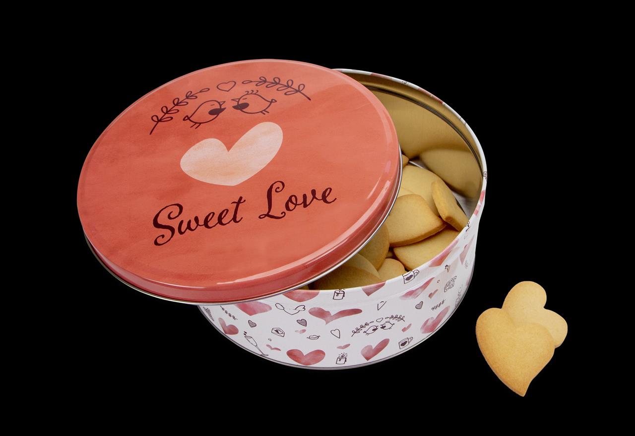 Die Abbildung zeigt eine runde Keksdose mit dem Aufdruck "Sweet Love" und einem Herz sowie zwei Vögeln auf dem Deckel. In der Dose befinden sich mehrere herzförmige Kekse.