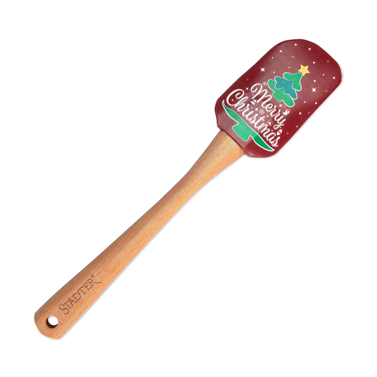 Dies ist ein Teigschaber mit einem weihnachtlichen Motiv. Auf dem roten Schaber steht „Merry Christmas“ und es ist ein grüner Tannenbaum abgebildet.