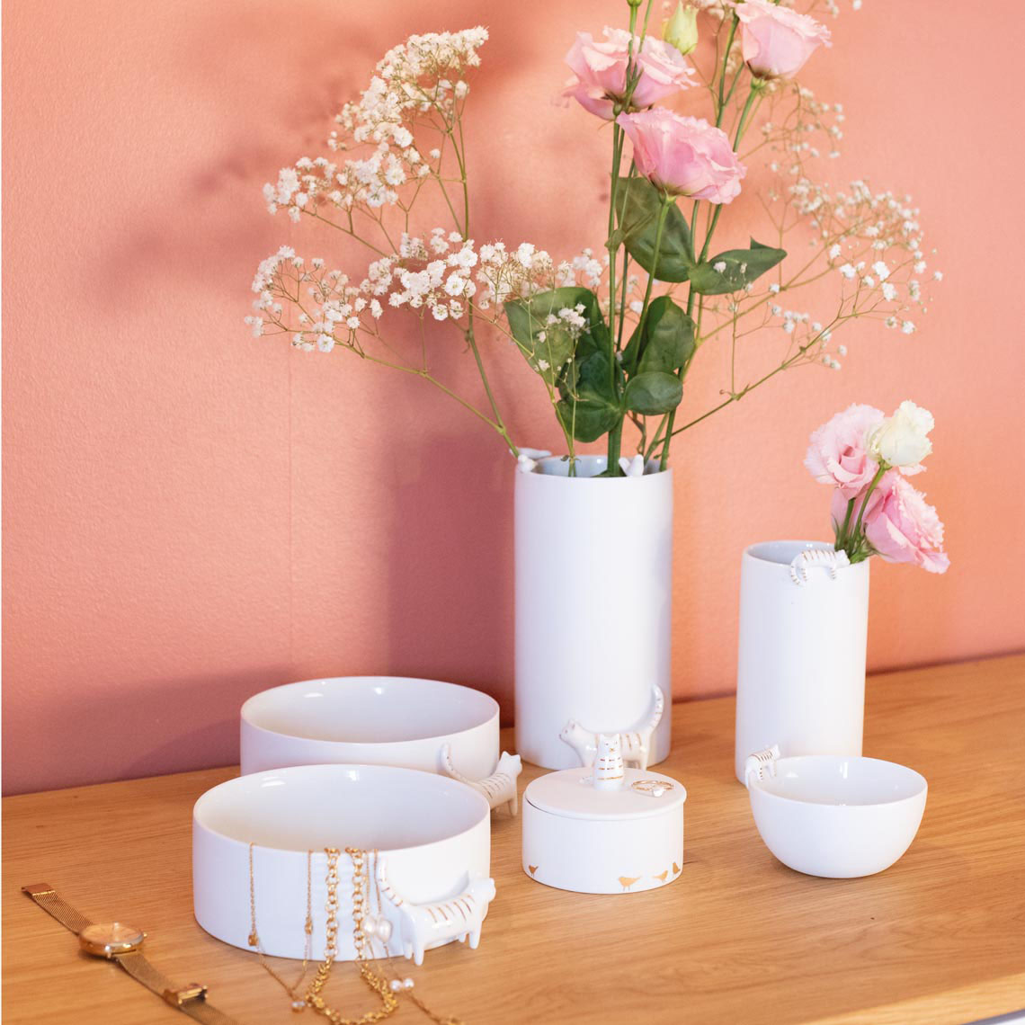 Auf dem Bild sind weiße Keramikschalen und Vasen auf einer Holzoberfläche zu sehen, die mit Schmuck und Blumen dekoriert sind. Im Hintergrund ist eine rosa Wand, die dem Arrangement eine warme Atmosphäre verleiht.